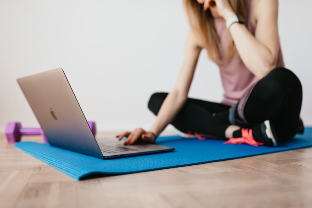 Crop young female in activewear browsing laptop while sitting on yoga mat on carpet